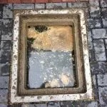 An outdoor drain with its cover removed, showing stagnant water and fat-like deposits indicating a blockage.